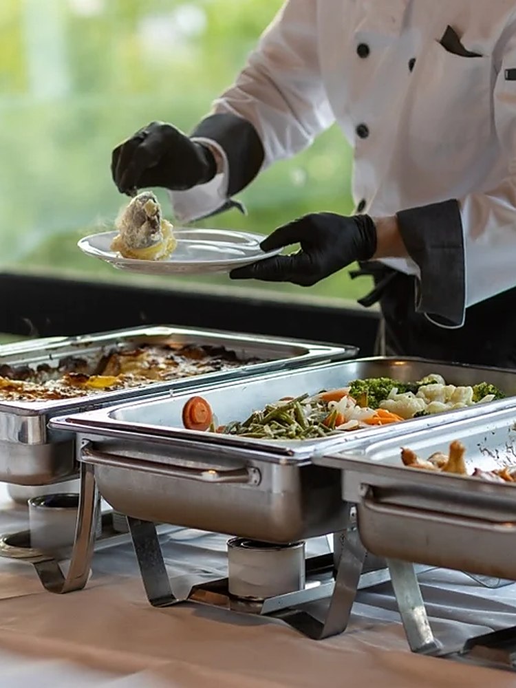 A chef in whites and serving gloves plating a portion from a row of chafing dishes at a buffet