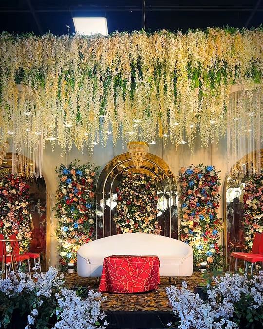 Wedding stage with a red lounge, framed by a canopy of hanging white florals and a flower-lined aisle